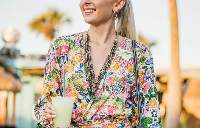 A girl in a brightly-printed dress and holding a margarita looks off to the side