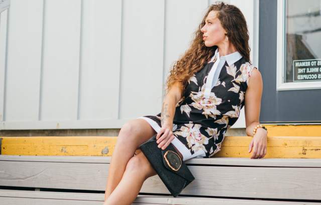 A smiling woman with long curly hair wears a black collared dress and sits on a step.