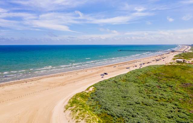 Drone image of a beach that shows the water, beach, and sand dunes from very high. Down the beach, a few cars and a pier are visible.