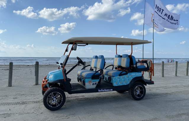 A light blue and patterned golf cart with two large flags sits on the beach.