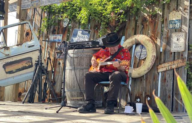 An elderly man in a red Hawaiian shirt and black cowboy hat strums a guitar on a wooden stage