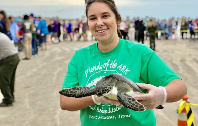A woman in a green shirt stands on the beach and holds up a sea turtle