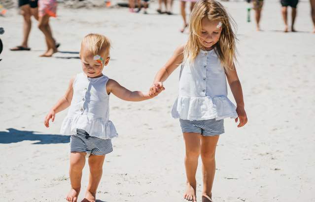 Two toddlers hold hands on the beach in matching outfits. Both of their faces are painted.