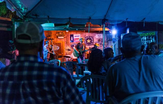 A dim crowd watches a brightly-lit singer wearing a straw cowboy hat and strumming a guitar