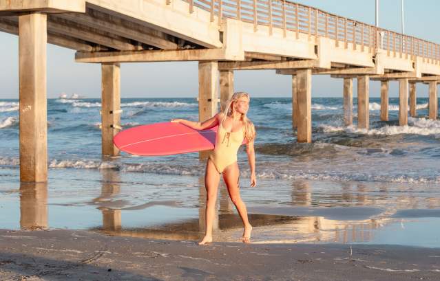 A woman in a yellow bikini carrying a red surfboard walks under a wooden pier.