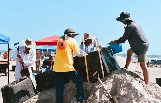 A group of four men crowd around forms on a pile of sand with buckets and sculpting tools