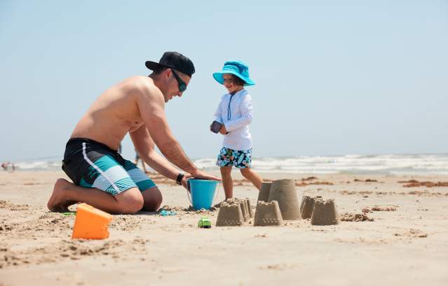 A man in a swimsuit kneels on the sand next to a child wearing sun gear and a hat. They pack sand in a blue bucket and build a sandcastle.