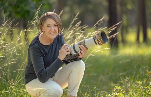A woman smiles while kneeling in a field with a large camera