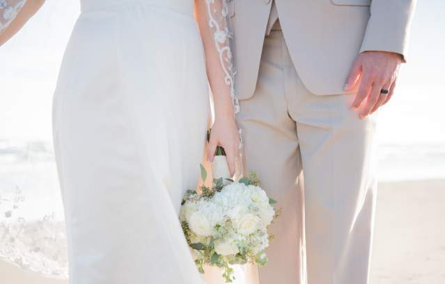A close-up photo of a couple standing on the beach in wedding attire. The bride holds a bouquet.