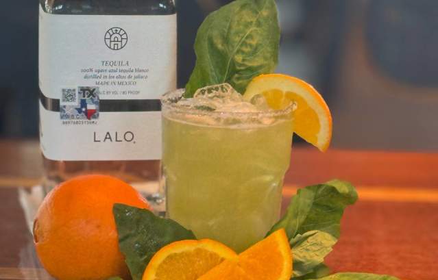 A light green margarita sits on a table surrounded by oranges and basils