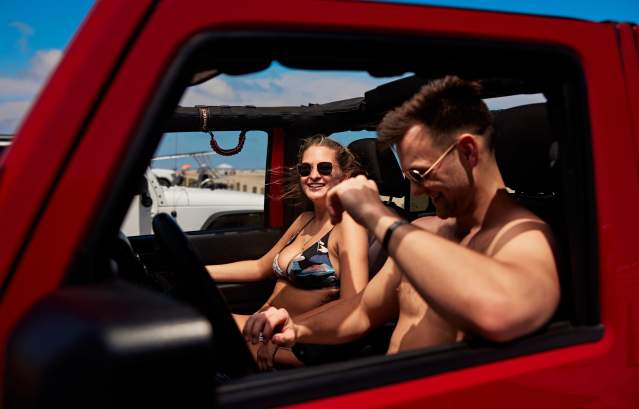 A man and woman in swimsuits are seen sitting in a red Jeep from the Jeep's window view