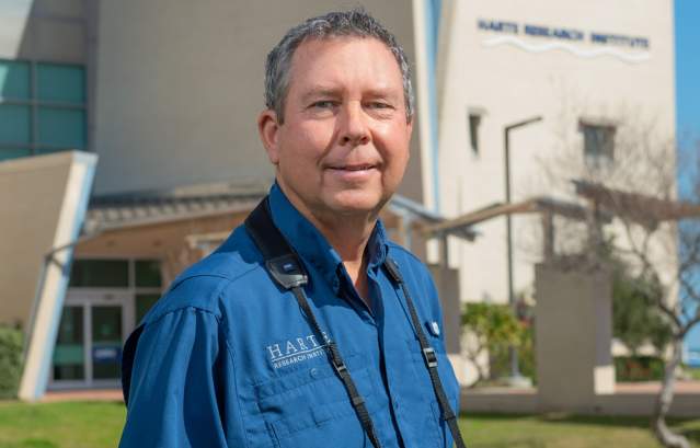 A man in a blue button up shirt smiles at the camera. He wears binoculars around his neck.