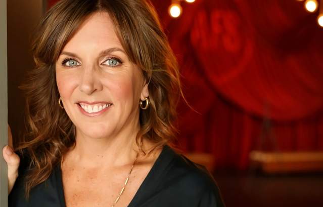 Portrait of Kathy Blackwell smiling in a black blouse with a warm, theatrical background.