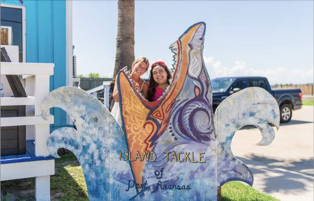 Two girls pose inside of a shark mouth cutout that reads "Island Tackle"