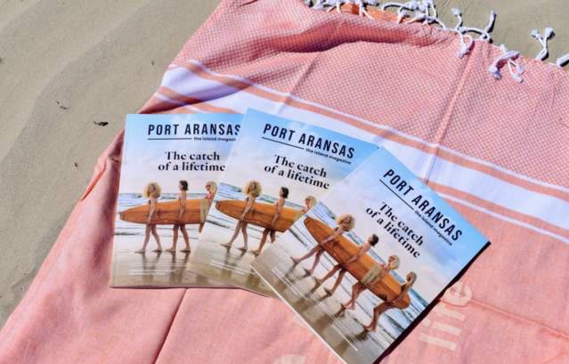 A group of three Port Aransas island magazines sits on an orange towel on the beach