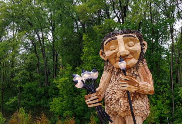 Large Wooden Troll sculpture smaelling lamps as if they were flowers against green trees.