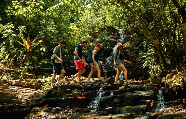 A group of hikers crossing a stream near Pozo Azul Waterfall, surrounded by many trees and nature.