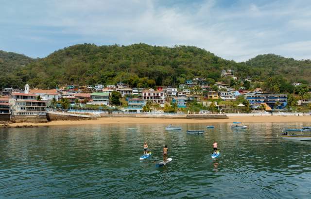 Three people practice paddleboarding in calm waters near the beach and colorful houses on the hillside of Taboga Island, surrounded by lush vegetation.