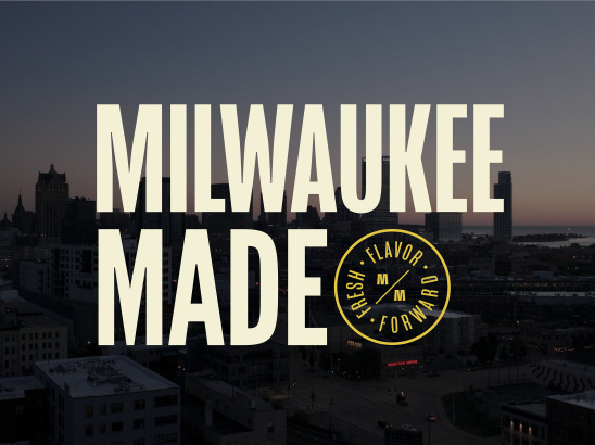 Aerial view of Milwaukee's downtown skyline at dusk with tall buildings silhouetted against a gradient sky. Large cream-colored text reads 'MILWAUKEE MADE' overlaid on the cityscape, with a circular yellow badge logo containing 'FRESH FLAVOR FORWARD' and stylized 'MM' letters positioned to the right of 'MADE'.
