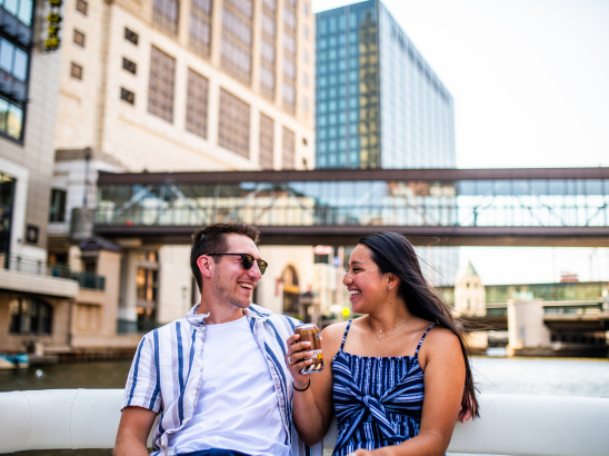 Couple laughing with each other on the back of a boat on the Milwaukee River with tall buildings in the background