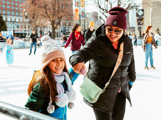 a little girl ice skating on a public ice rink while holding her mom's hand