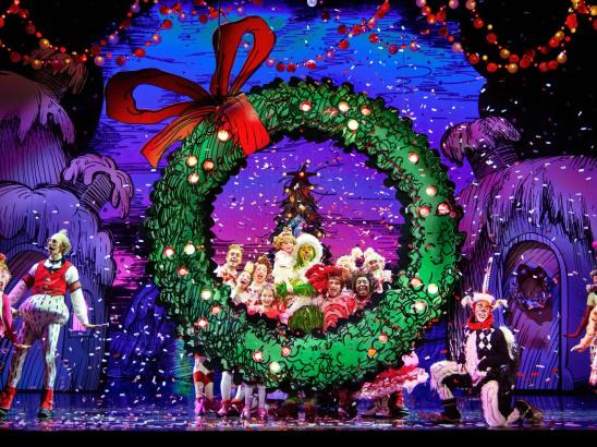 A colorful stage scene from a Christmas-themed musical featuring characters in bright costumes gathered around a giant green wreath with a red bow. Snow-like confetti falls from above, and whimsical, snow-covered houses are visible in the background.