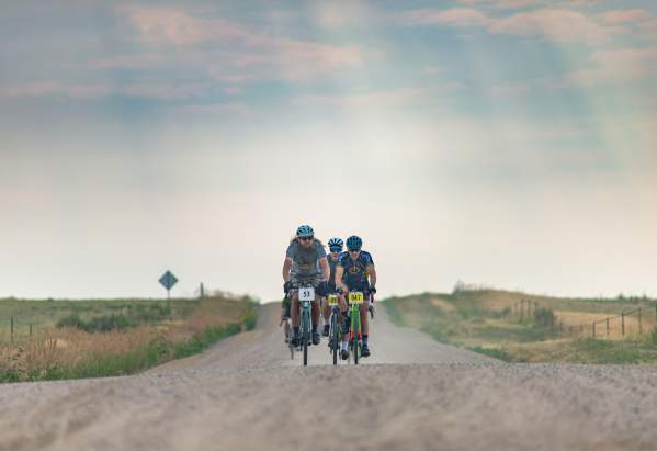 A group of cyclists ride along a gravel road, surrounded by open fields and a hazy sky in Fort Collins