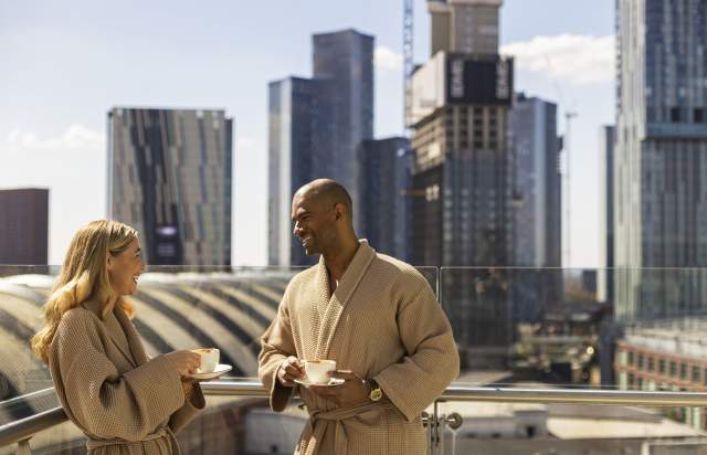 Couple on hotel balcony in dressign gowns drinking coffee with views over Manchester