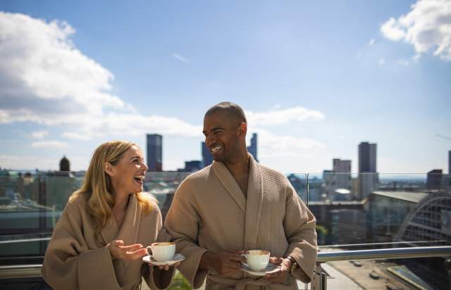 Couple in dressing gowns with cups of coffee stood on hotel balcony overlooking Manchester