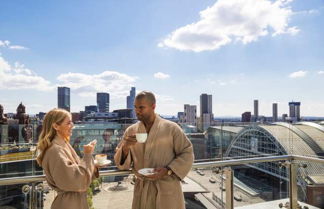 Couple in dressing gowns drinking coffee on Hotel Balcony overlooking Manchester