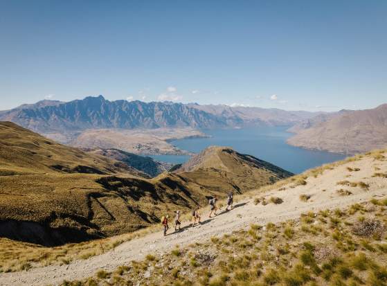 A group of hikers climbing the top of a mountain with views of Lake Whakatipu and Remarkable Mountains in the background