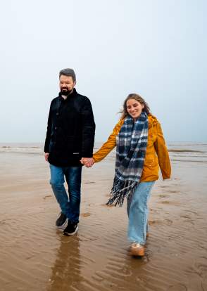 A couple wearing coats and scarves walk along a sandy beach