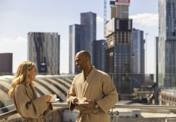 Couple on hotel balcony in dressign gowns drinking coffee with views over Manchester