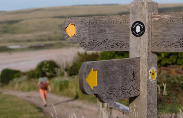 A wooden sign post with yellow arrows pointing to England coast path, with a walker in the distance