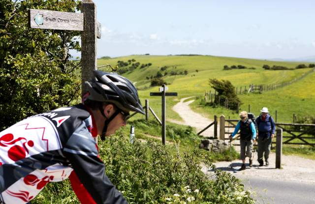 A mountain biker rides along the South Downs Way, with walkers on a trail in the background