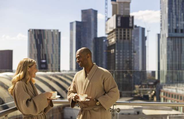 Couple on hotel balcony in dressign gowns drinking coffee with views over Manchester