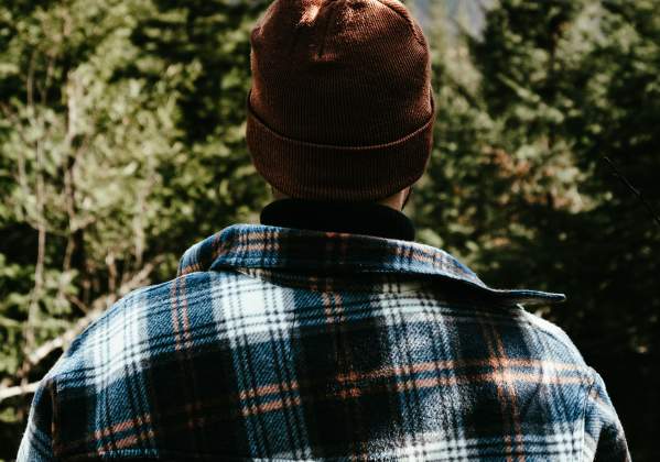 A person in a plaid flannel shirt and knit cap looks toward forested mountains.