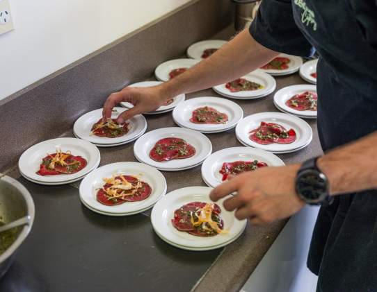 A person carefully plating small servings of food onto multiple white dishes arranged on a countertop, with a bowl of green sauce nearby