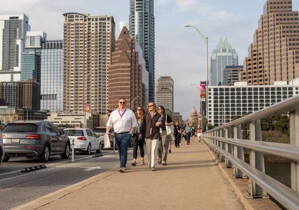 A group of people wearing SXSW badges walk across the Congress Avenue Bridge with the Austin skyline in the background.