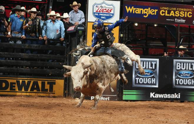 Bull rider on bucking bull with team behind gate in the background.