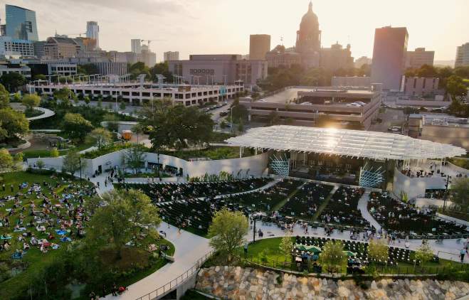 Aerial view of Moody Amphitheater in evening sun. There is a crowd on the open lawn and in seats near the pavilion stage. The State Capitol building is visible behind the amphitheater