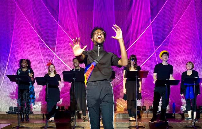photo of a performance at Trinity Street Playhouse. A man wearing black athletic clothing stands in the center, singing, and a chorus stands behind him. Each chorus member is singing and stands behind a black music stand