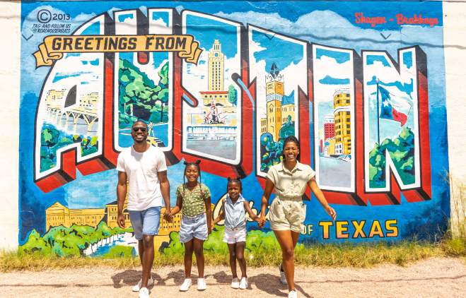Family of four with two young daughters hold hands and smile at camera in front of postcard mural that reads Greetings from Austin Texas