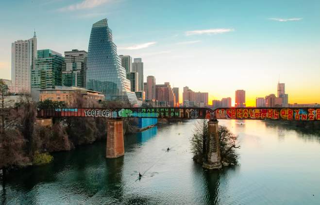 Kayakers paddling under a bridge with grafitti on Lady Bird Lake while the sun sets against Downtown Austin buildings.