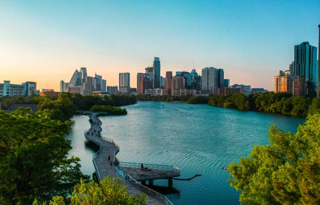 People walking along suspended board walk that follows the shape of the Colorado River, leading to Downtown Austin skyline.