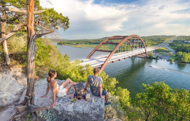 Hikers sitting on a hilltop rock overlooking the Pennybacker Bridge at Lake Austin.