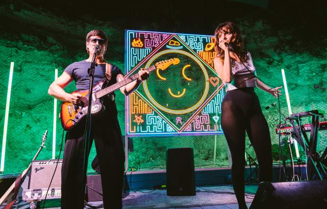 Man playing guitar and woman singing into a microphone on a stage backlit by a neon smiley face.