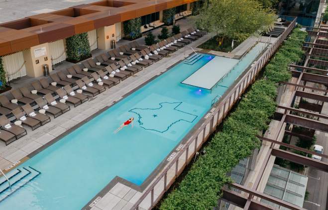 Aerial photo of the JW Marriott rooftop pool with a woman in a red bathing suit swimming on her back in the center of the pool. The floor of the pool includes an outline of the state of Texas with a star on the location of Austin.