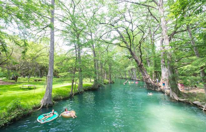 People floating on Blue Hole in pink and blue innertubes, with trees lining the shoreline.