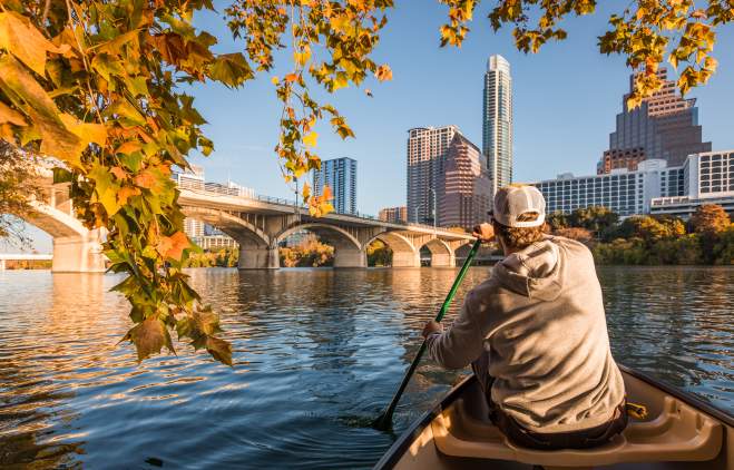Man in Canoe on Lady Bird Lake across from downtown Austin Texas skyline with fall leaves